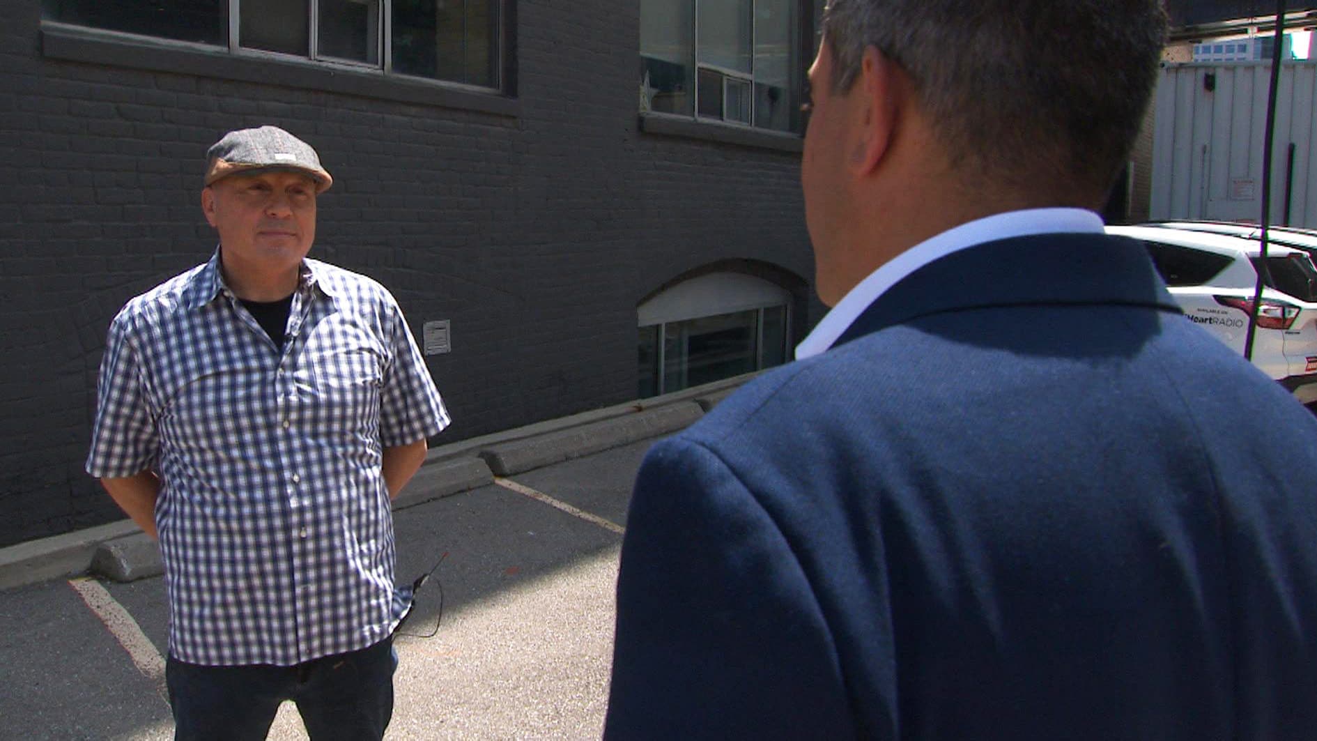 A man in a checkered shirt and a golf cap speaks to a reporter in a parking lot.