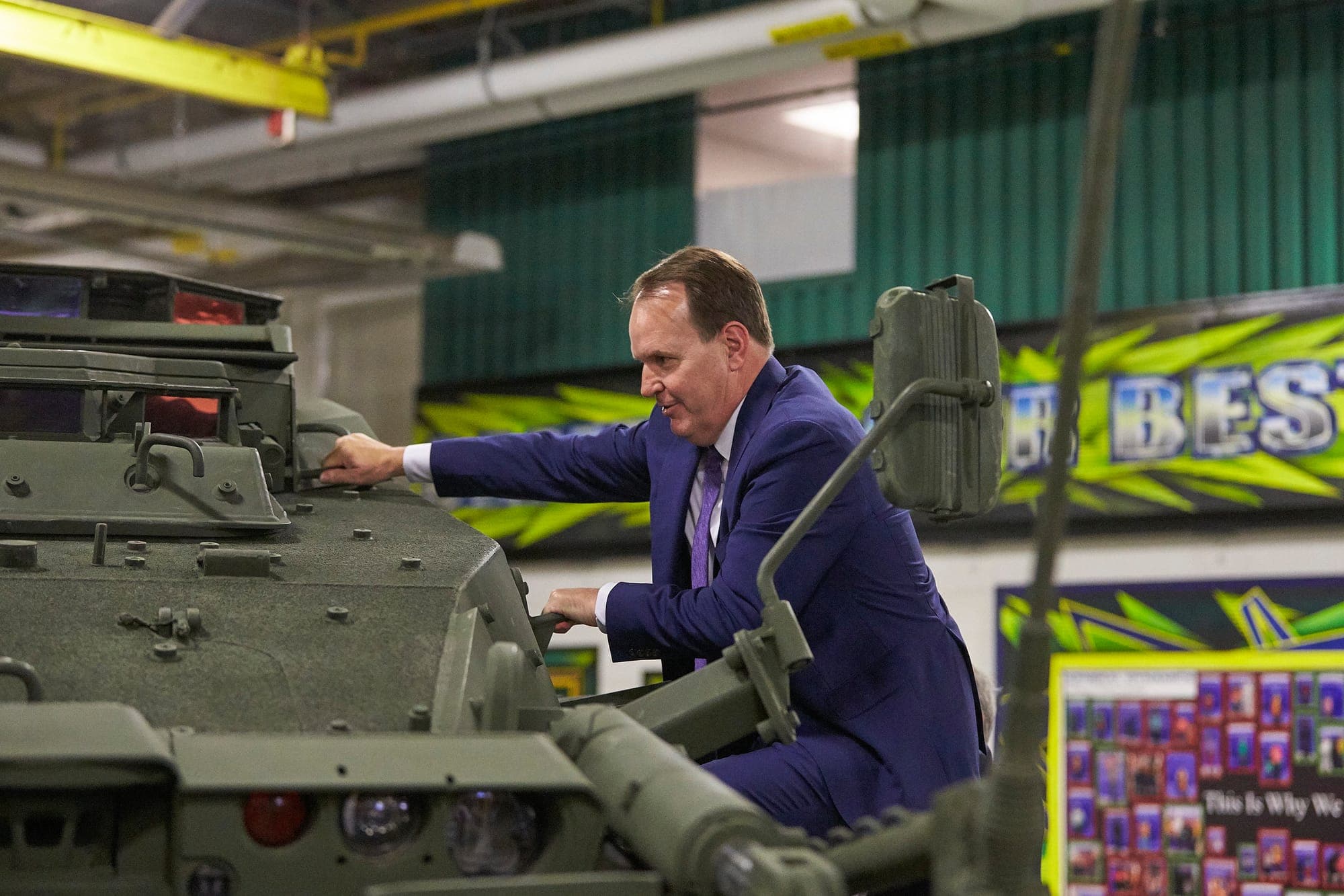 A man in a blue suit climbs into the side of an army-green armoured vehicle. 