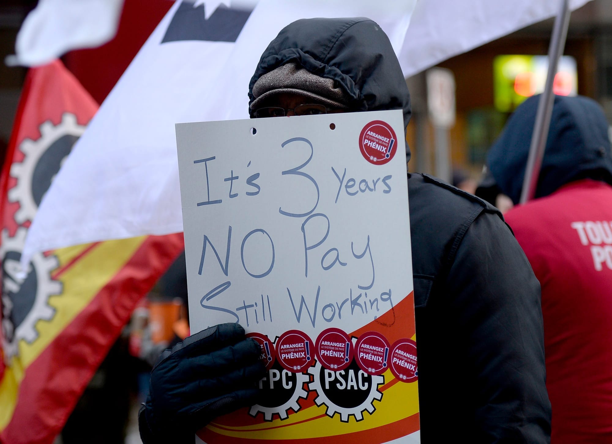 A person holds a protest sign reading "It's 3 years. No pay, still working."