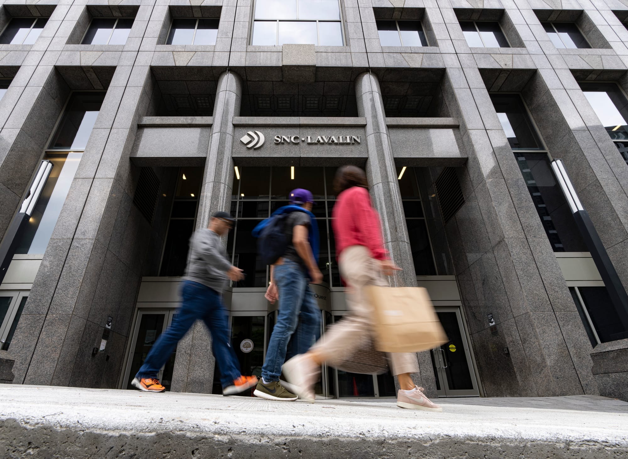 Three people walk past a building with a sign that says 'SNC-Lavalin.'