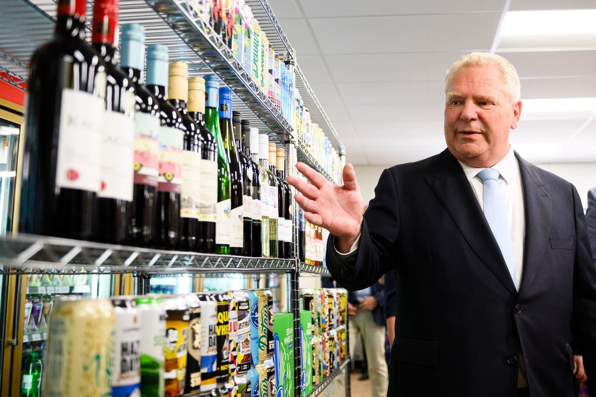 Ontario Premier Doug Ford wears a suit and tie and gestures to wine bottles on the shelf of a store.