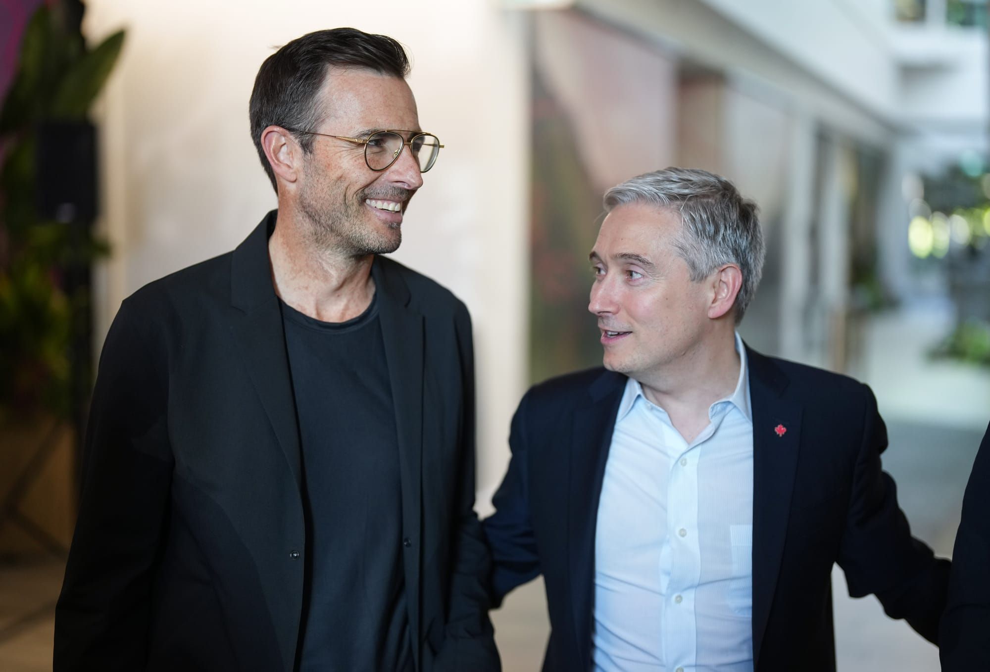 A man wearing a glasses and black jacket over a t-shirt stands next to a man wearing a suit and Canadian maple leaf pin. 