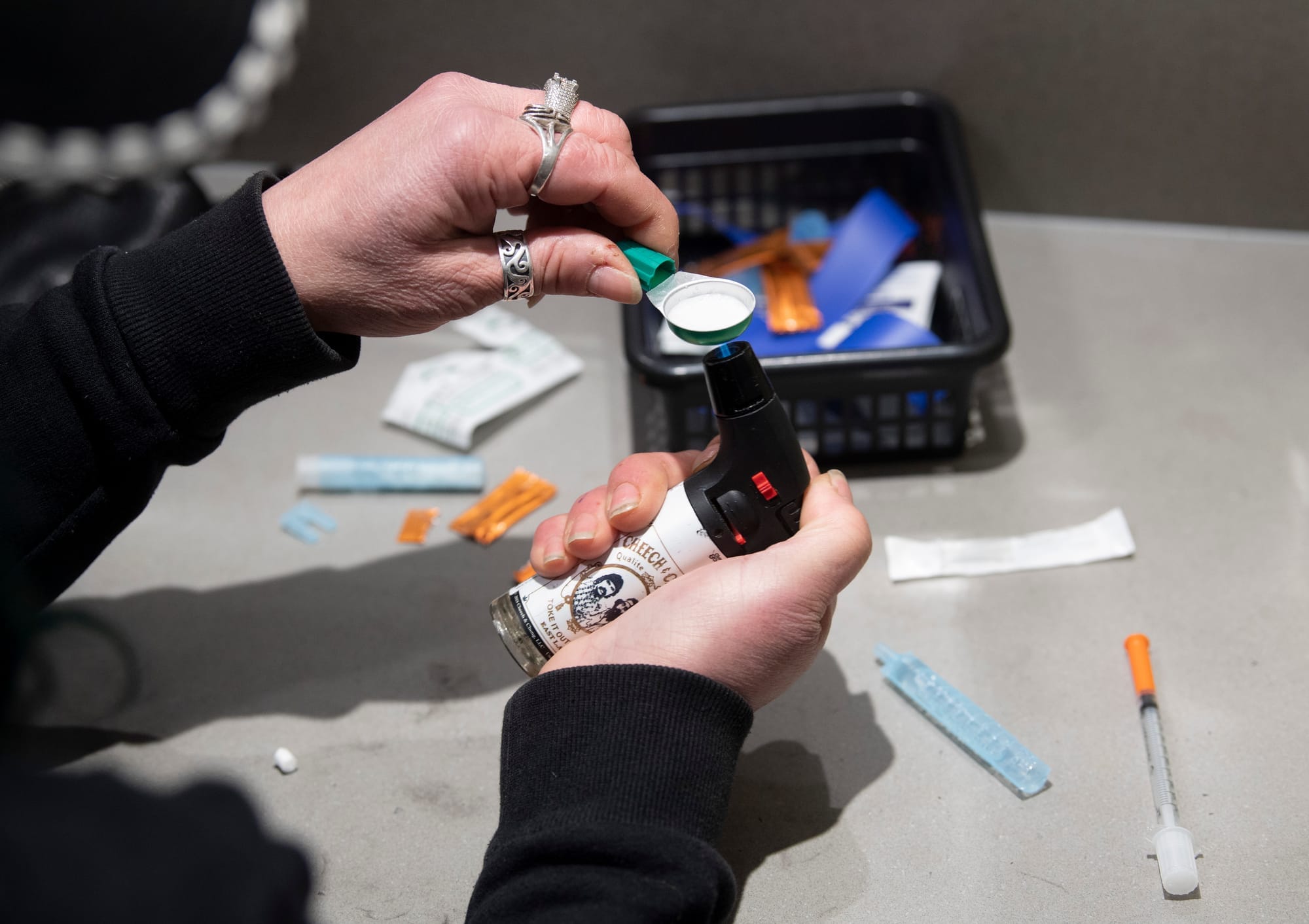 A pair of hands with chunky silver rings hold a drug-filled spoon over a butane lighter.