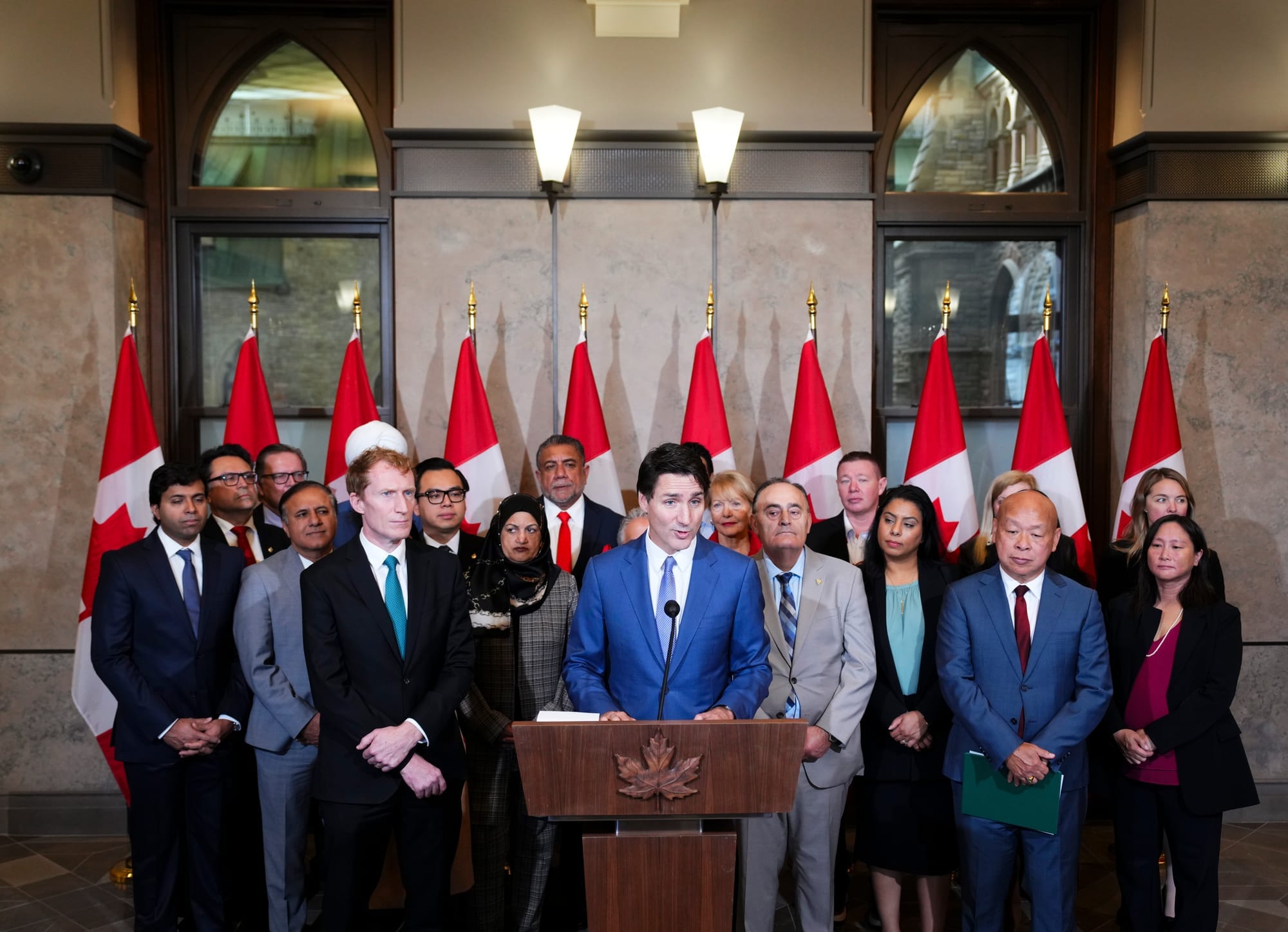 Prime Minister Justin Trudeau stands at a podium speaking while members of his cabinet stand behind him looking on. Cabinet m