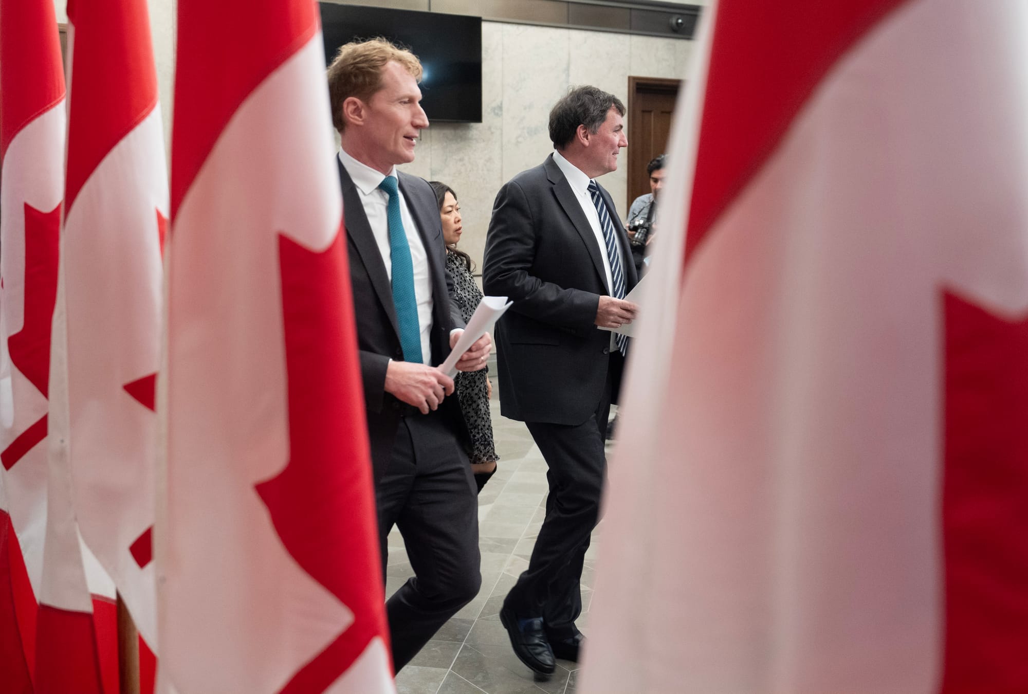 Three walking people are shown through a gap in a row of Canadian flags.