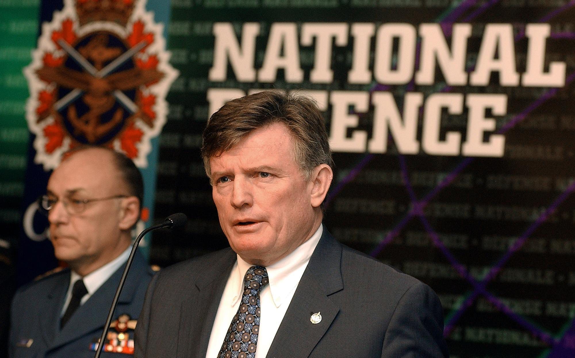 A man in a grey suit and patterned tie stands behind a microphone in front of a backdrop with the words NATIONAL DEFENCE