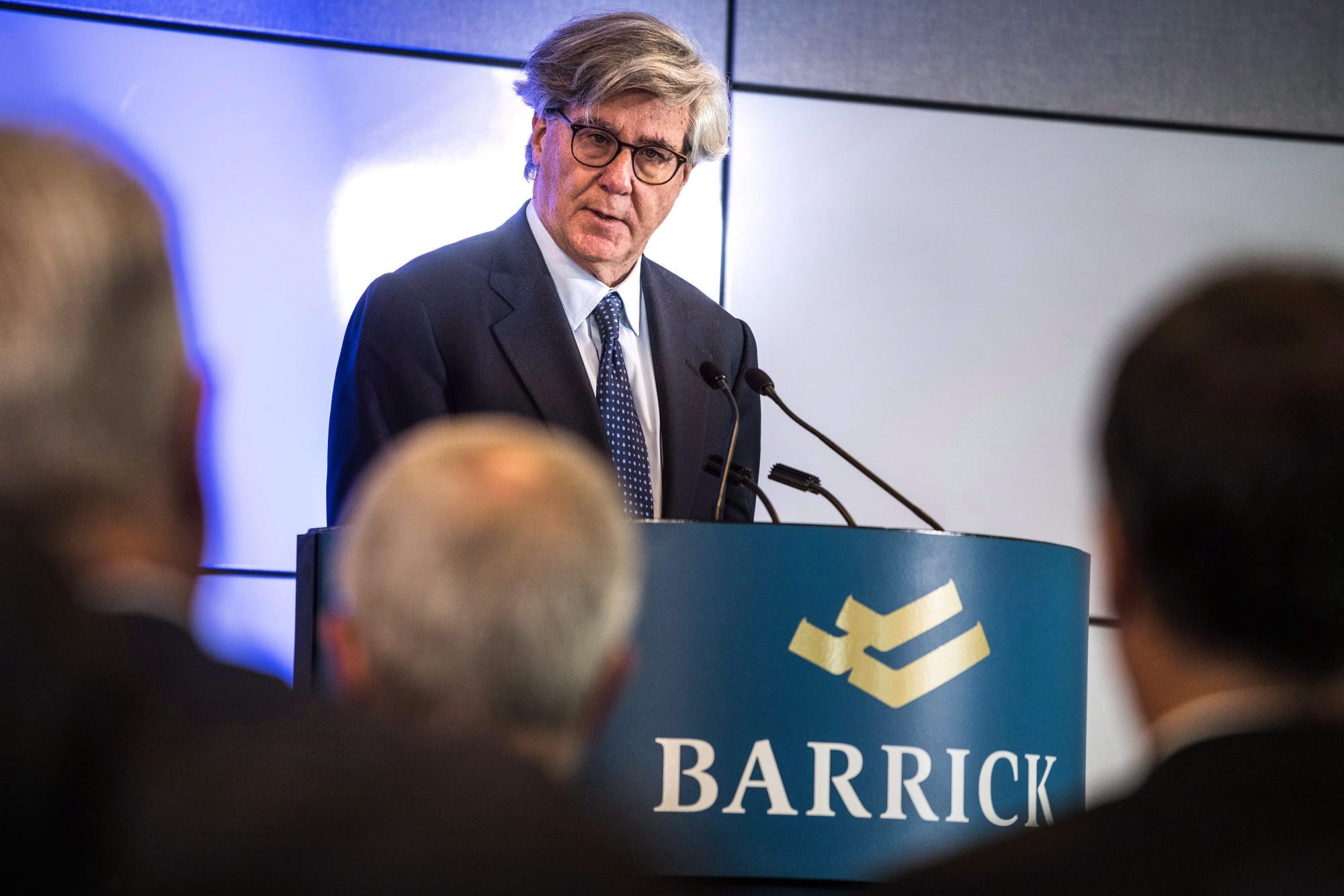 A grey-haired man in a suit and tie stands behind a podium featuring the word BARRICK