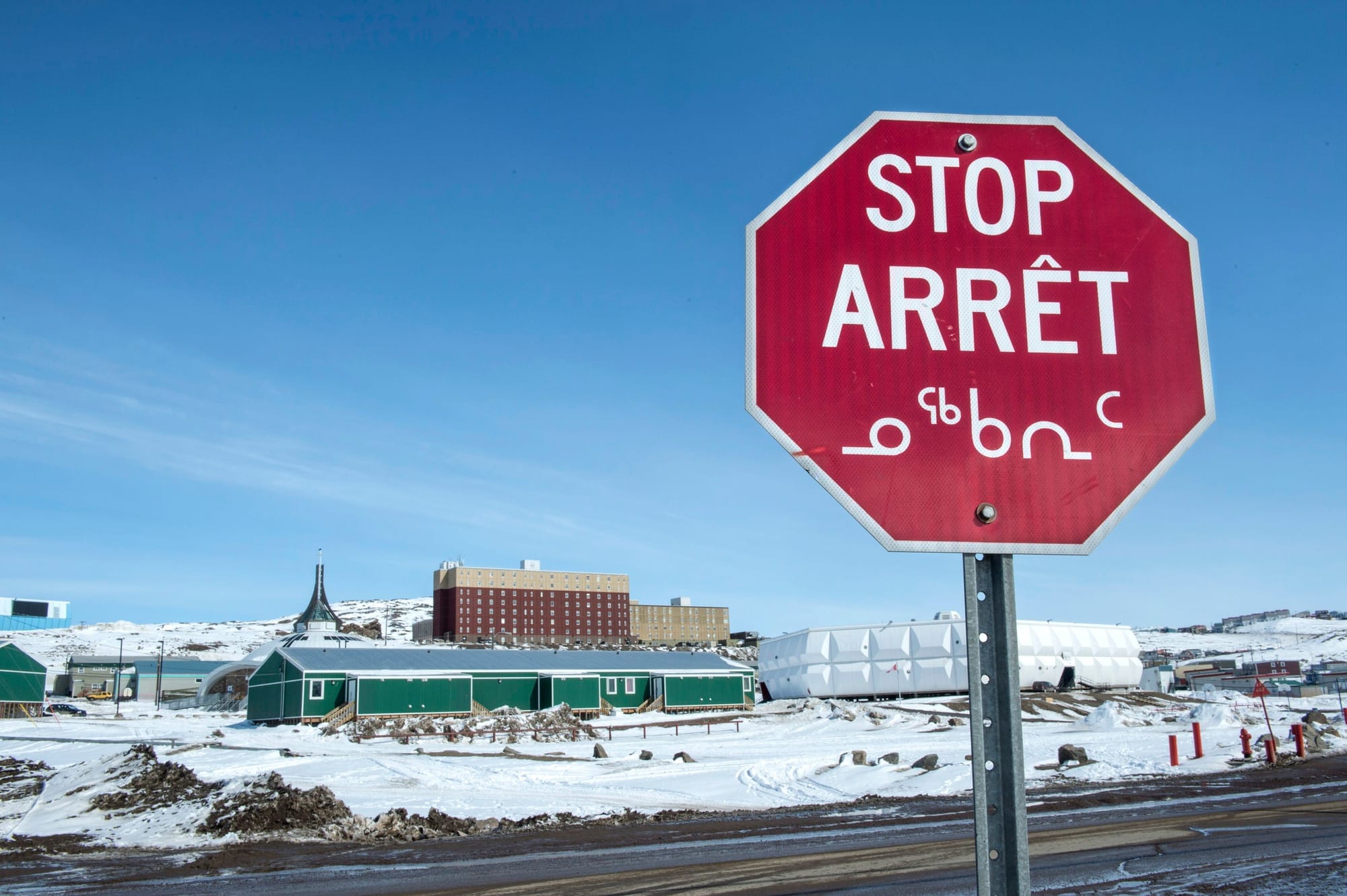 A stop sign featuring French and Inuktut is seen in front of buildings in a snowy scene in Iqaluit.