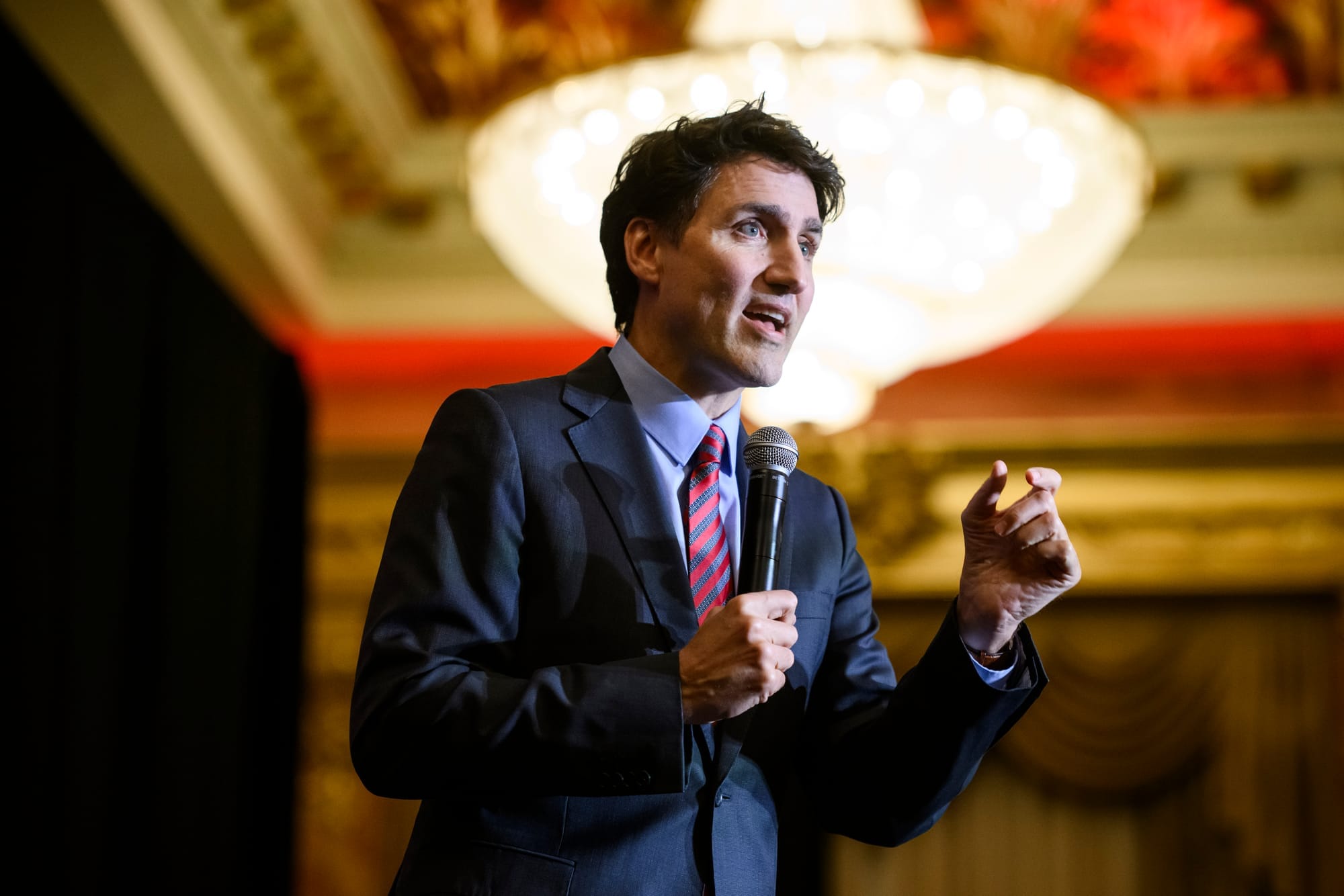 Justin Trudeau stands speaking holding a microphone. A chandelier and ornamented ceiling is visible behind him.