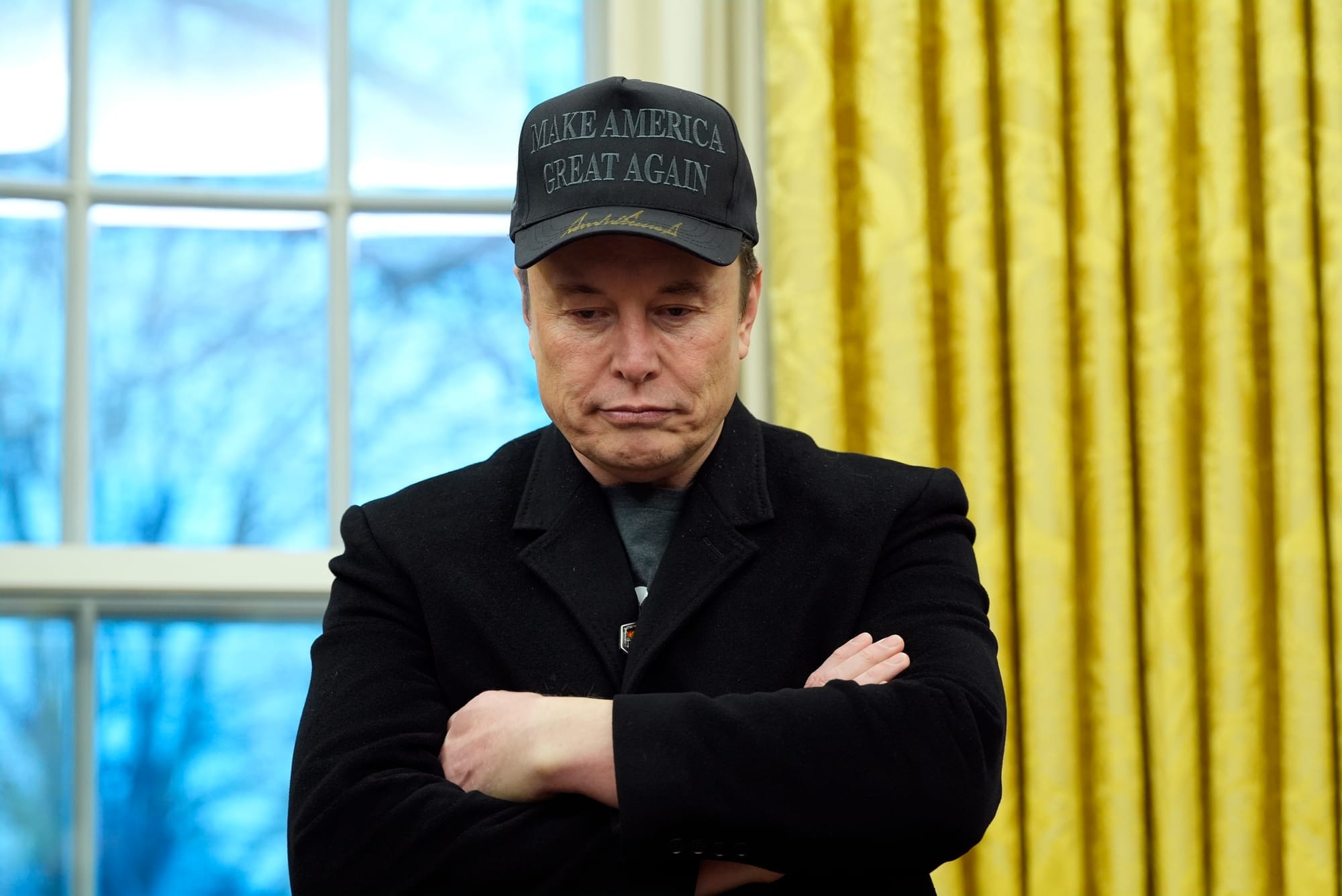 Elon Musk wears a black 'Make American Great Again' ballcap and a black blazer as he stands with his arms crossed.