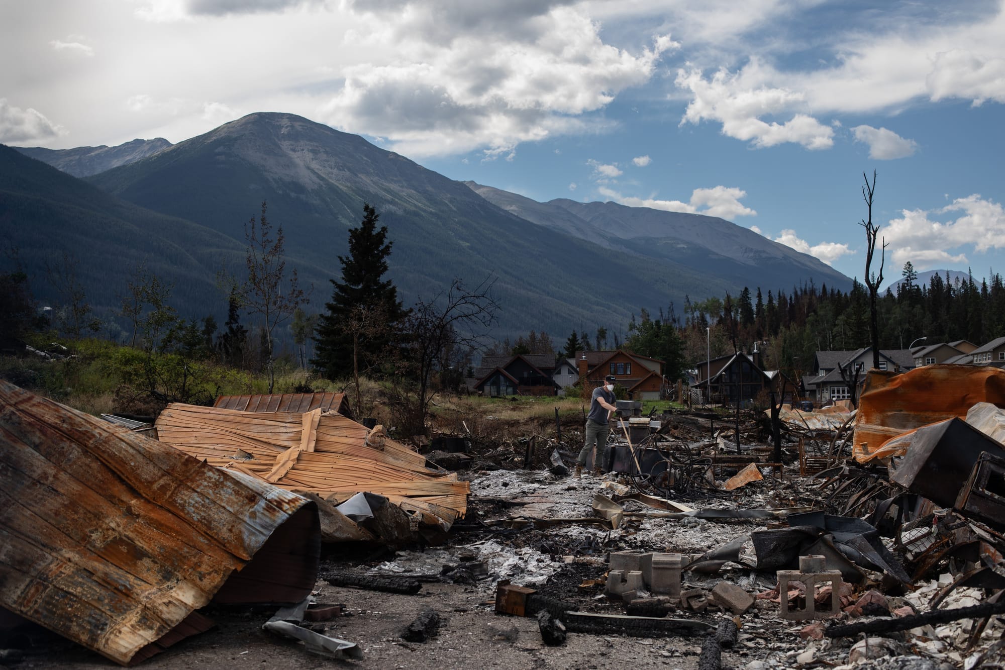 A homeowner wears a respirator mask and digs in burned rubble. A mountain and trees can be seen in the background.