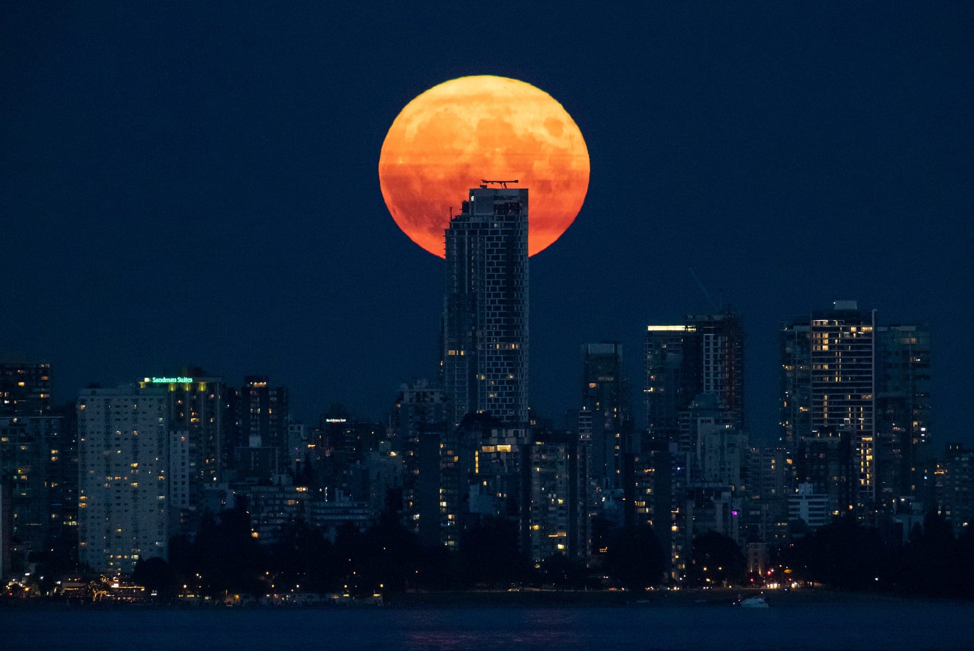 A bright orange full moon rises behind a condo tower in a nighttime photo of the downtown Vancouver skyline.