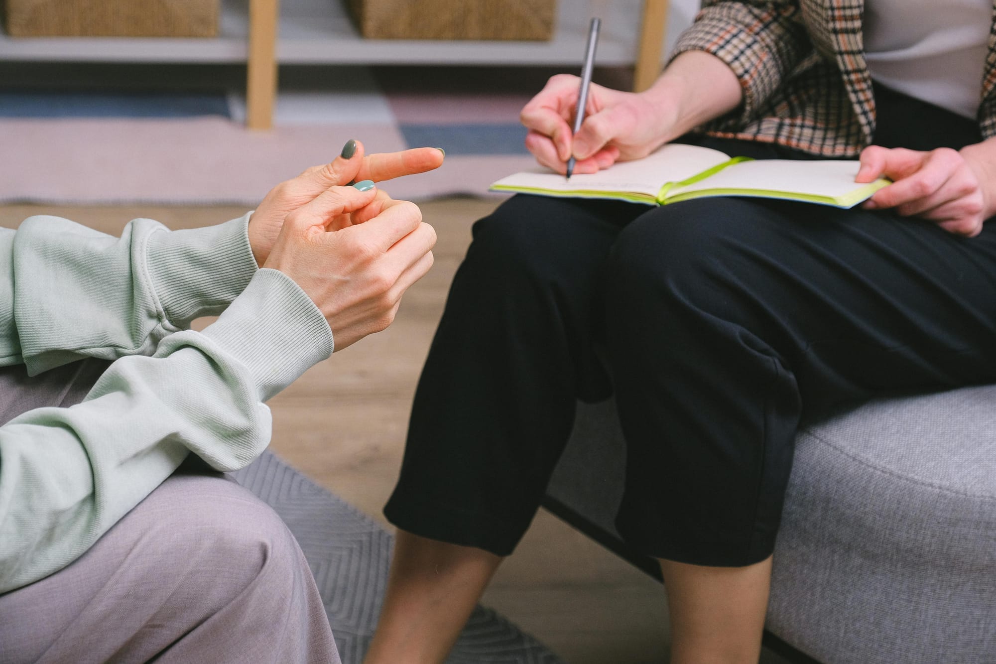 A person's hands are shown as they sit in a therapy office talking to a counsellor, writing information in a notebook.
