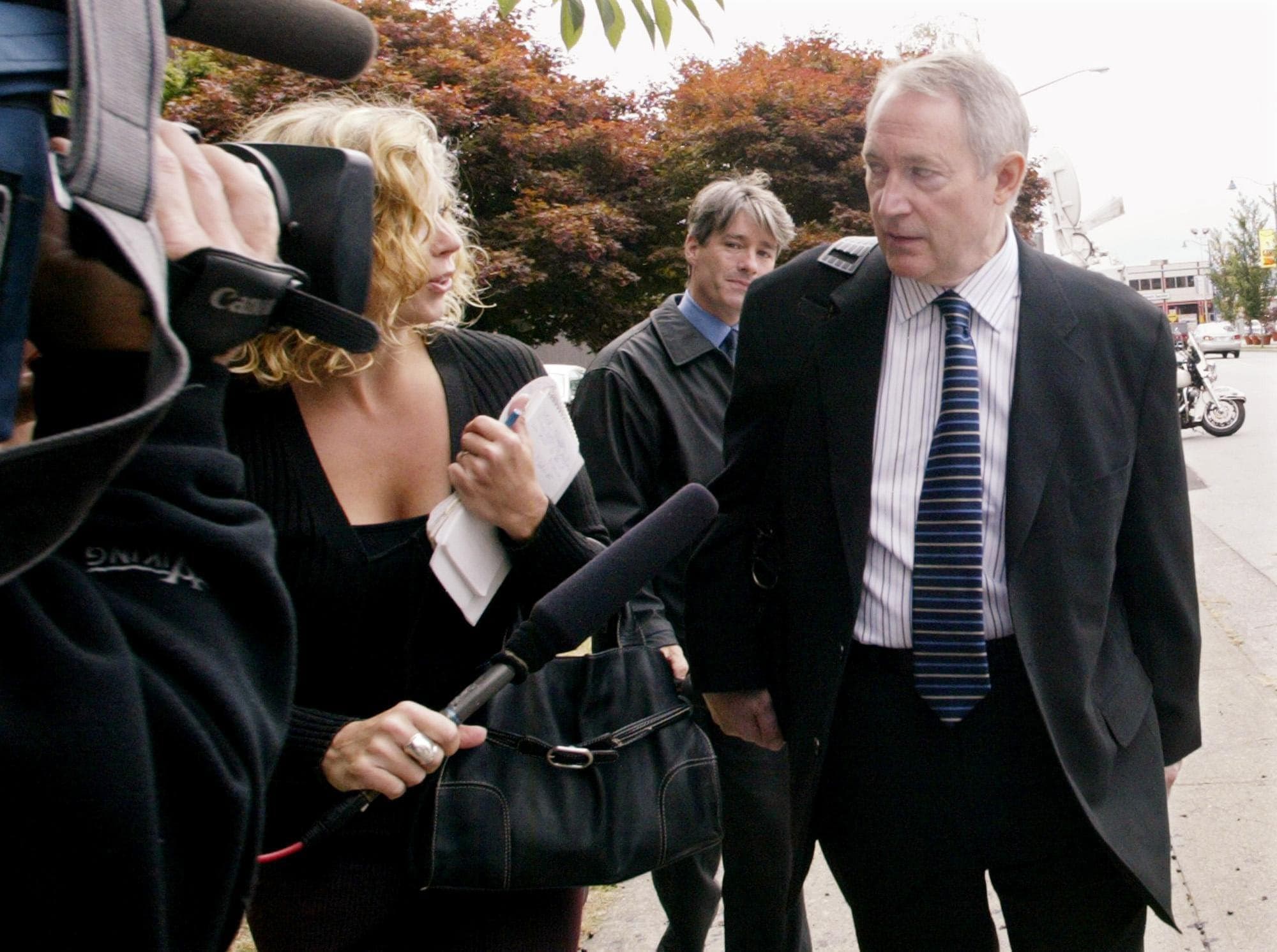 Michael Bolton, dressed in a black suit and striped tie, speaks to reporters on a sidewalk.