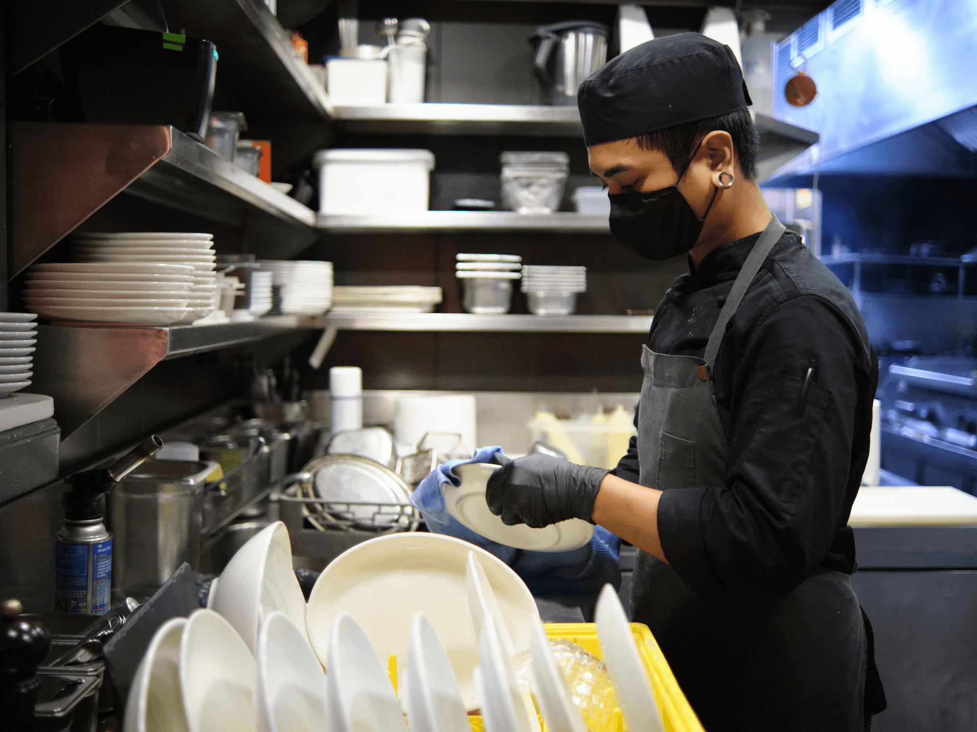 Close-up of a worker wearing protective mask and gloves cleaning dishes in a kitchen.