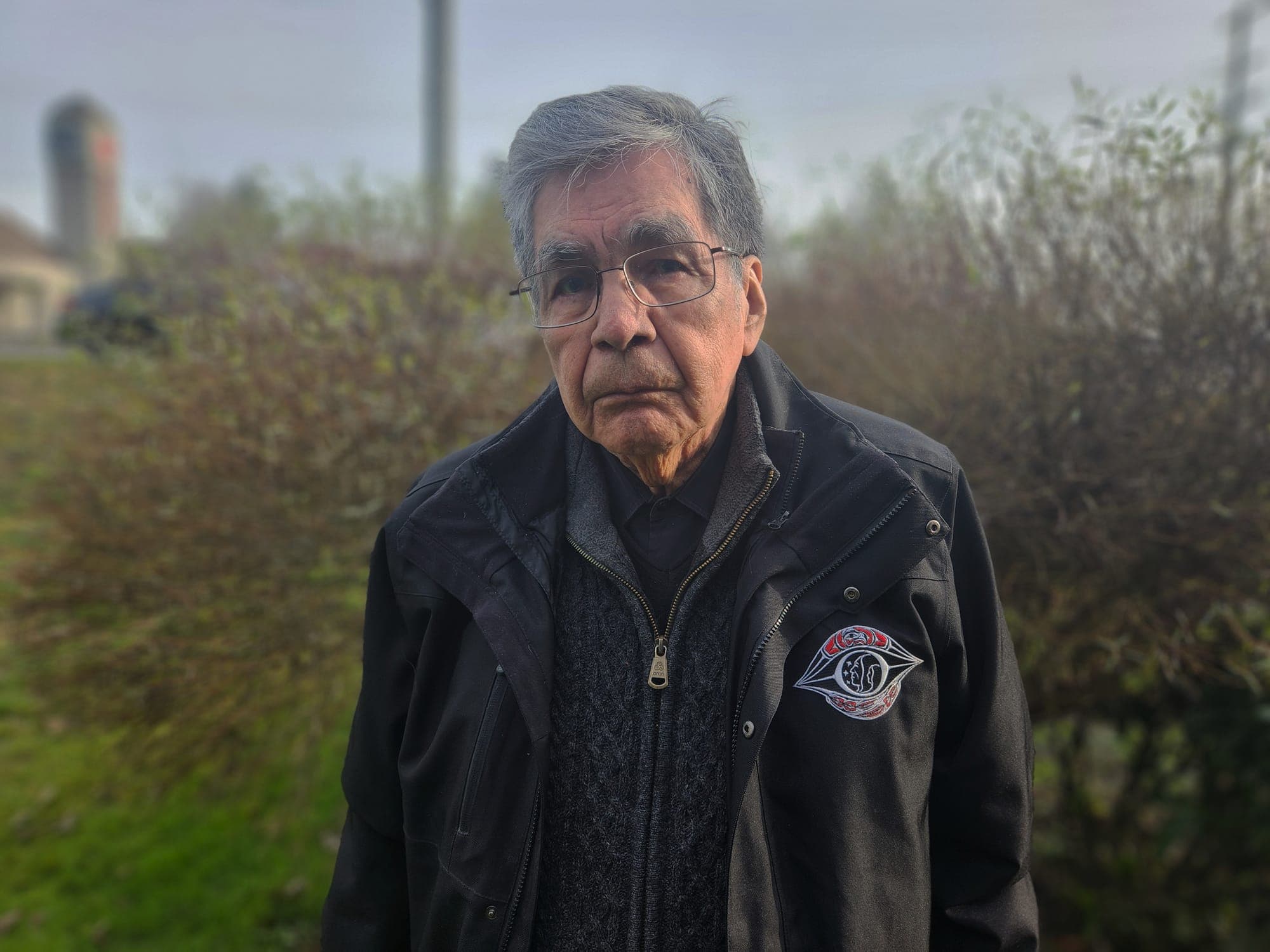 Joe Norris looks directly into the camera. He is an Indigenous man with grey hair and glasses, wearing a black jacket.