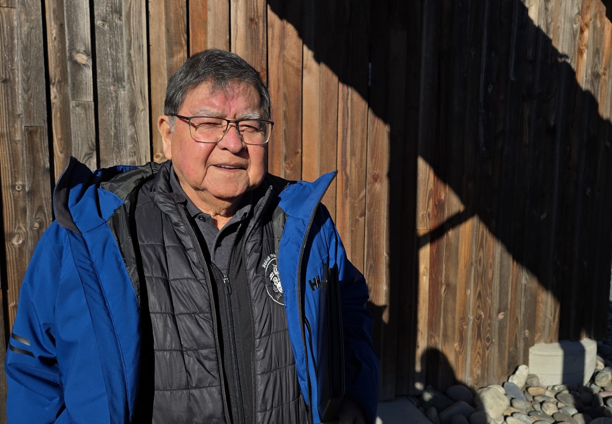 Clarence Pennier wears a blue coat with a dark-coloured lining underneath. He has short grey hair and glasses, and squints in the bright sun.