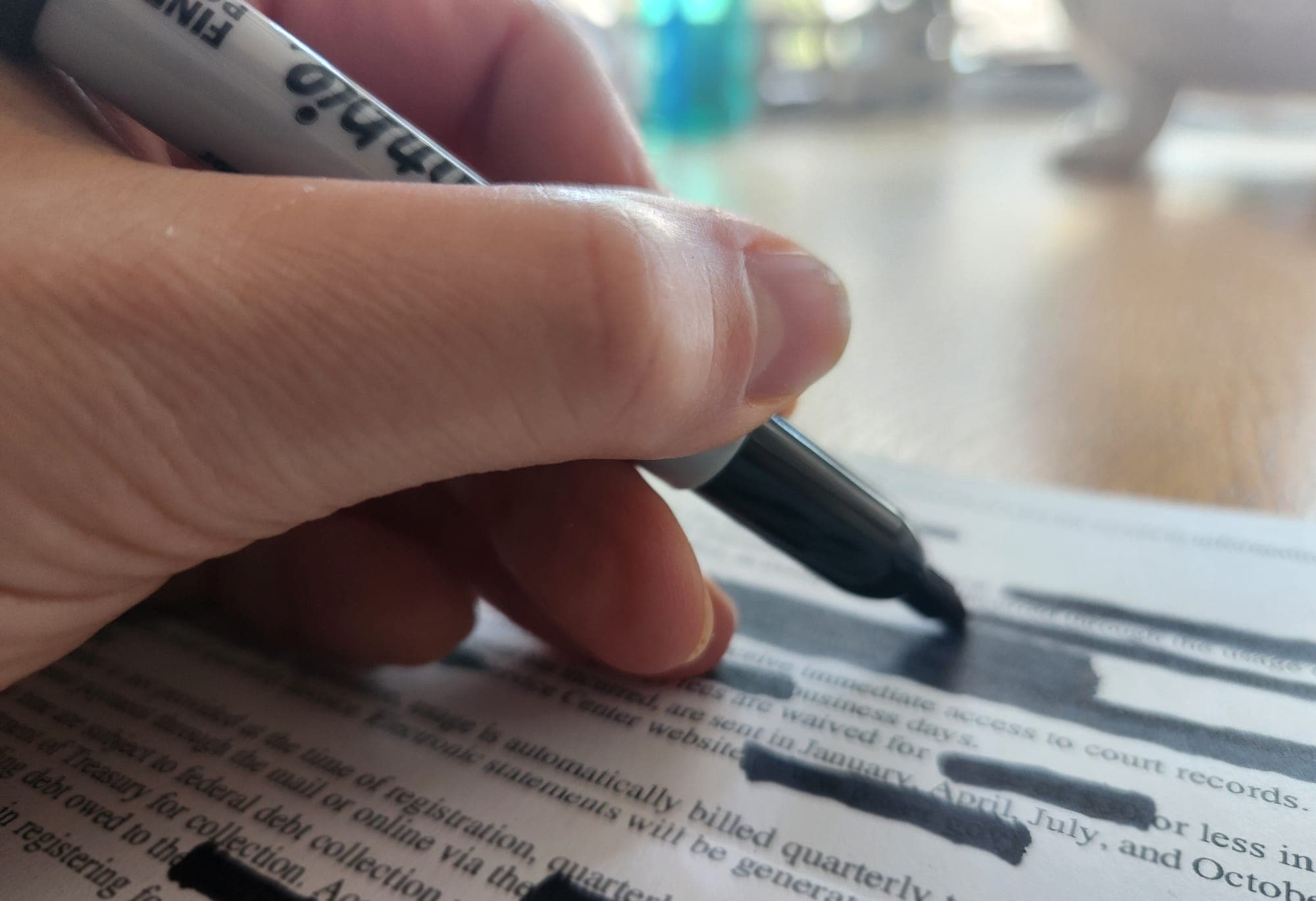 A hand holds a black marker over a document with numerous blacked-out lines of text.