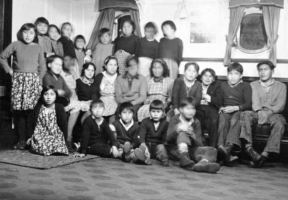 A black and white photo shows about two dozen Indigenous children posing together inside a room in a residential school. Some of their faces are blurred because of movement.