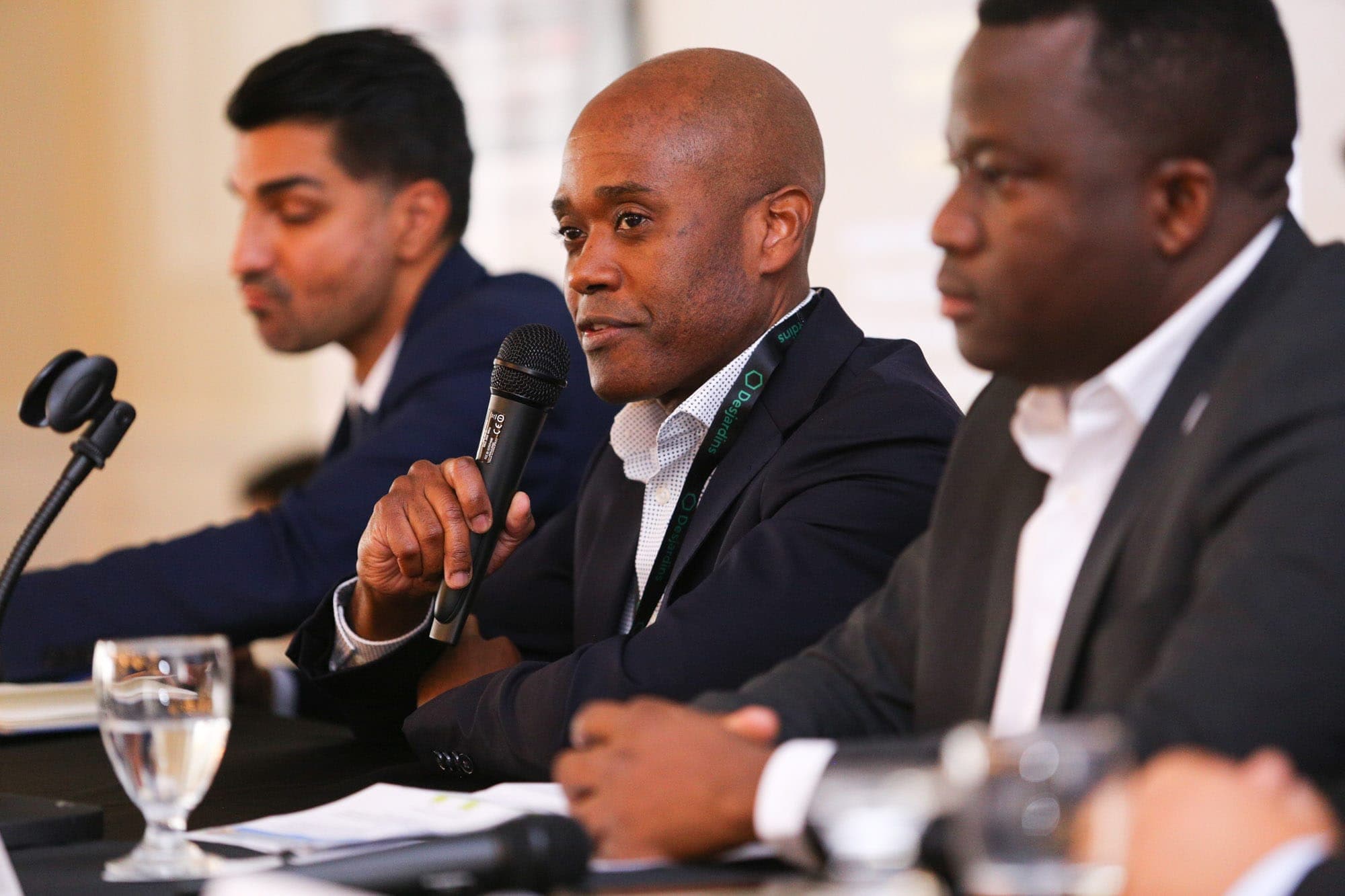 Johnson Joseph, a bald Black man in a dark suit, sits between two other men. He holds a microphone and speak during a panel.