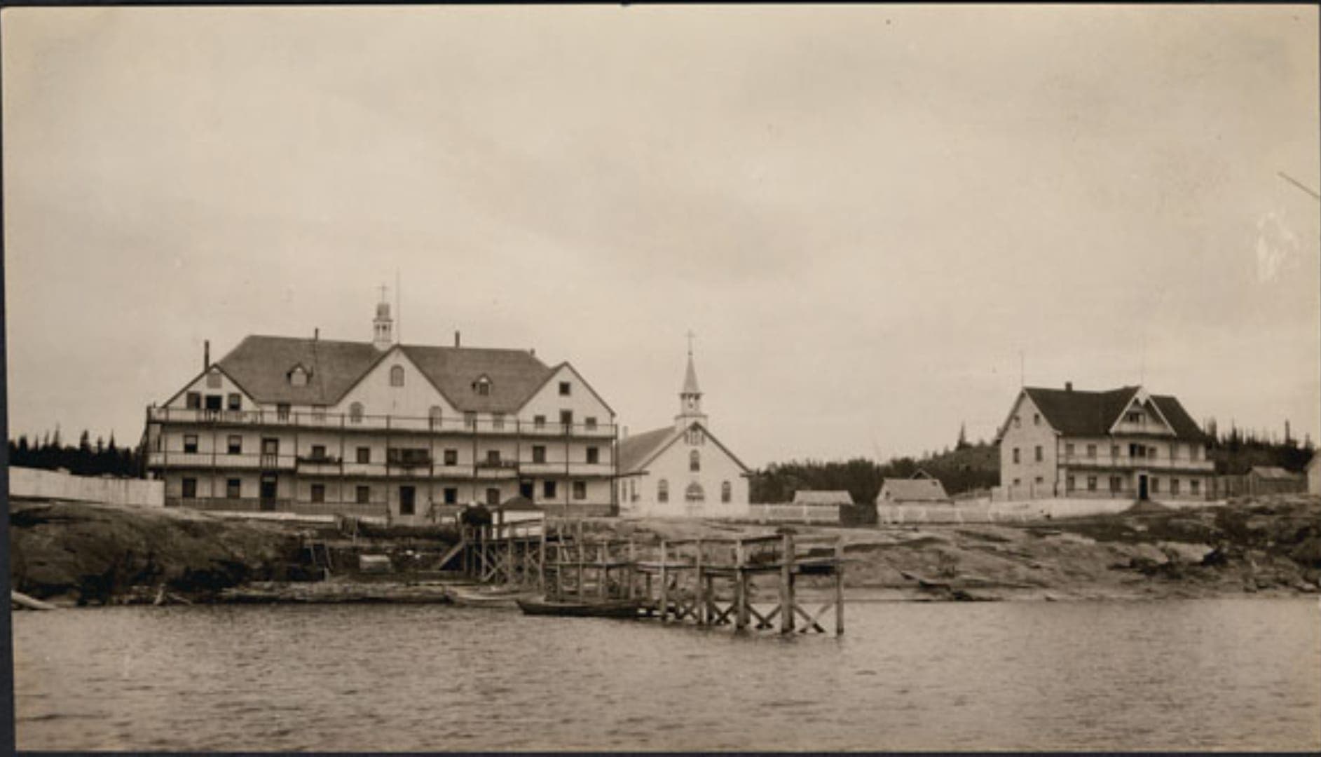 A sepia-toned black and white photo shows a residential school beside a chapel, with a dock leading into the water.