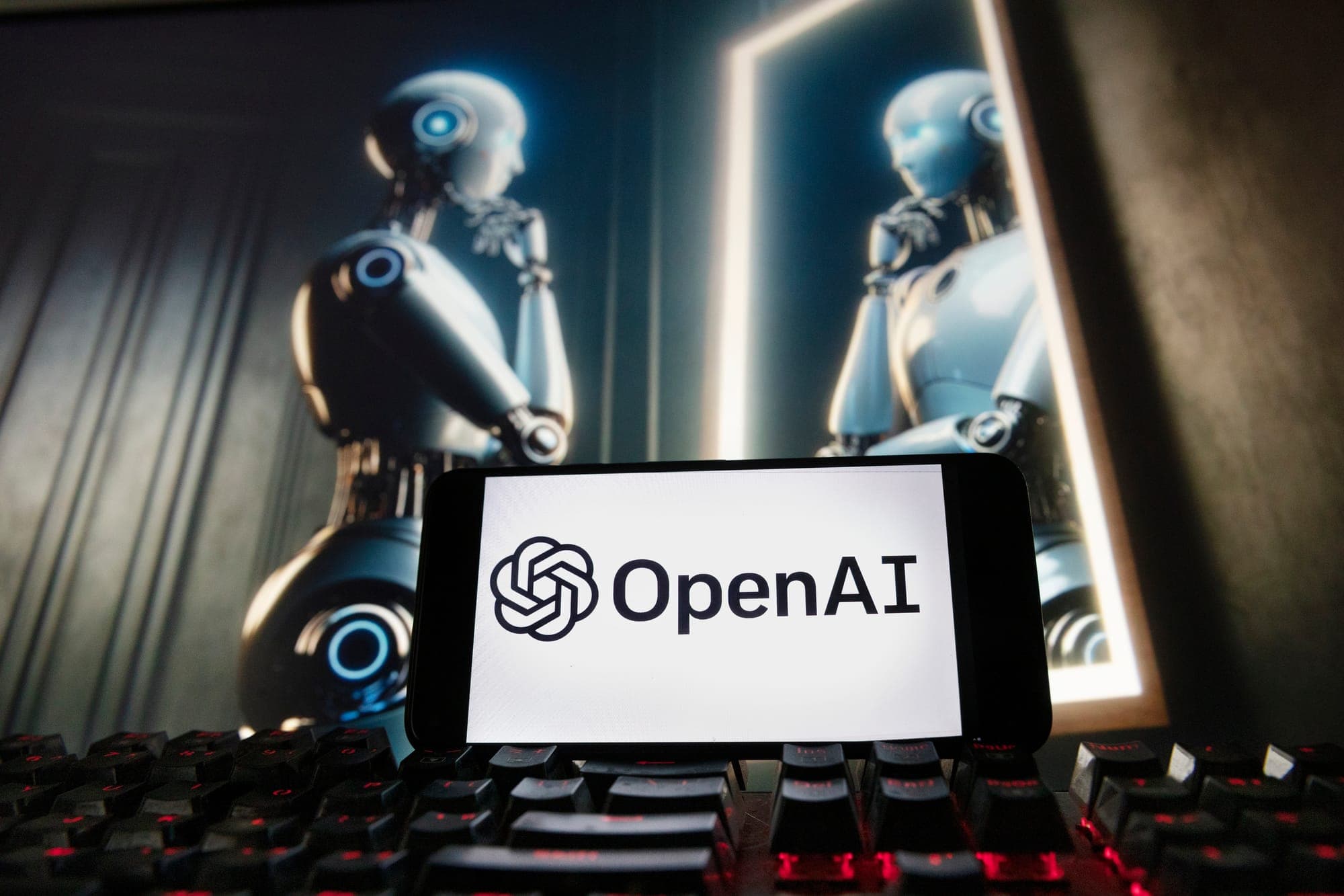 The OpenAI logo is displayed on a cell phone with an AI-generated image of a robot looking in the mirror in the background