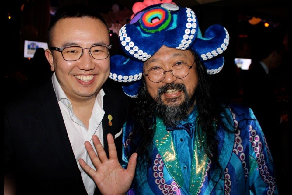 Jerry Zhong, an East Asian man in glasses, a white shirt and a black suit jacket, smiles as he poses next to Takashi Murakami, who is wearing a blue octopus hat and suit.