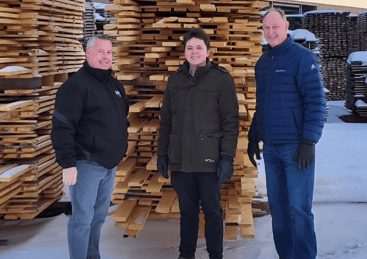 Three men in winter coats stand in front of pallets of lumber.