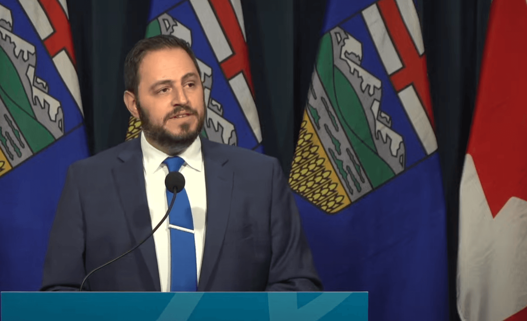A man in a suit stands in front of three Alberta flags and one Canadian flag.
