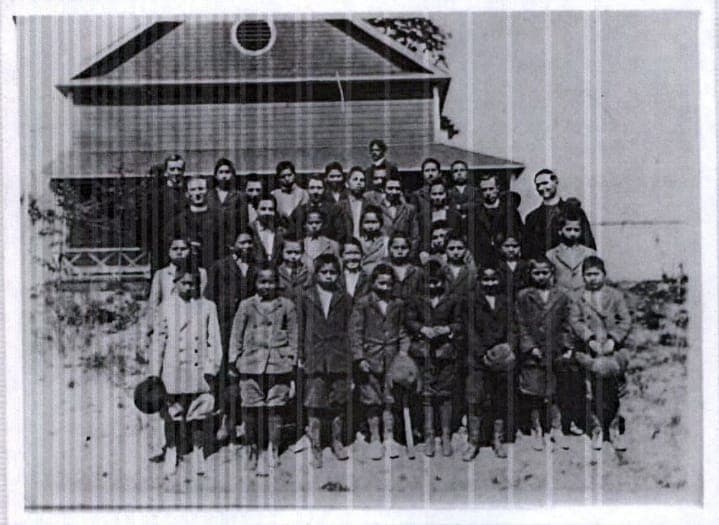 A group of Indigenous children wearing dark suits pose next to two men in clerical collars in a black and white photo.