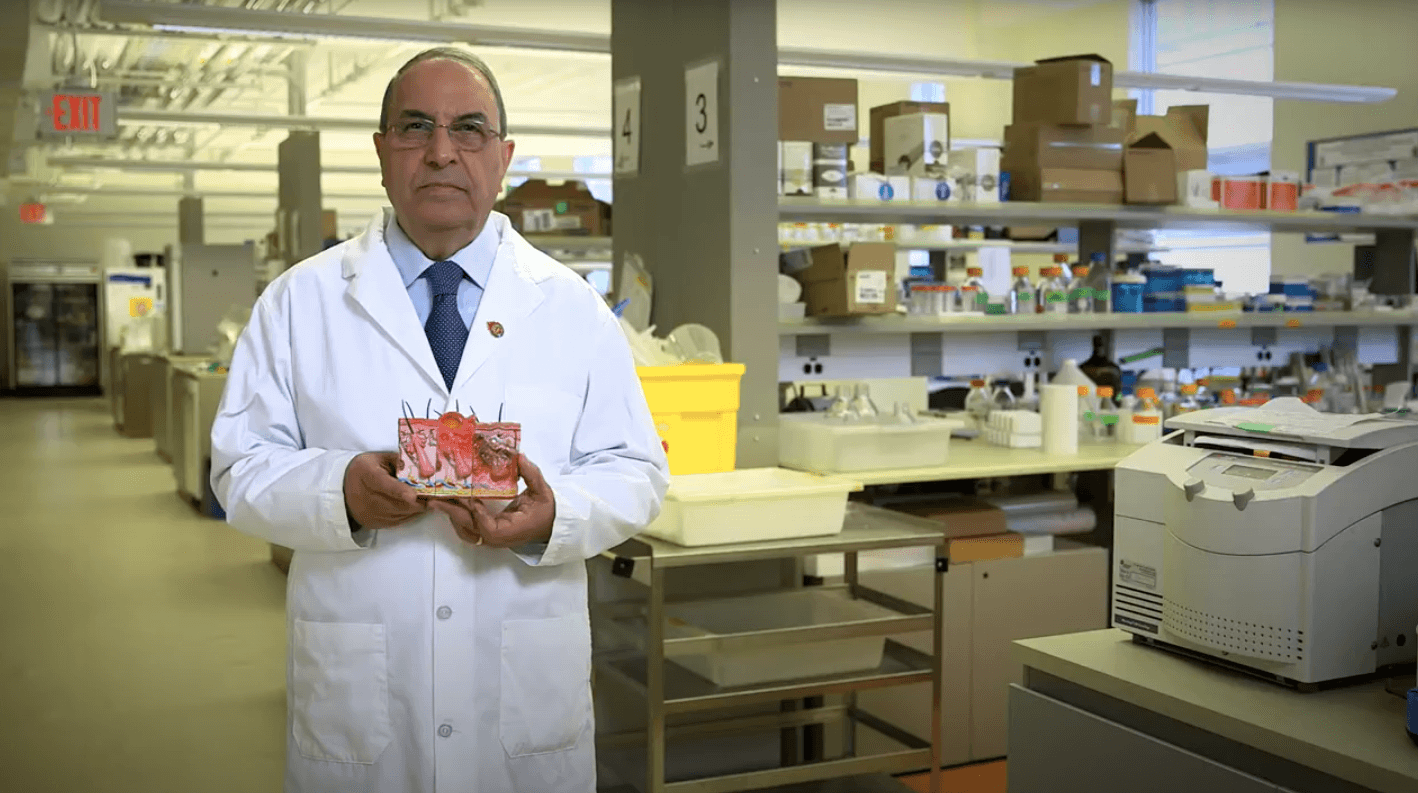 Aziz Ghahary, an older Persian man, stands in a lab wearing a lab coat and holding a model of a cross-section of human skin.