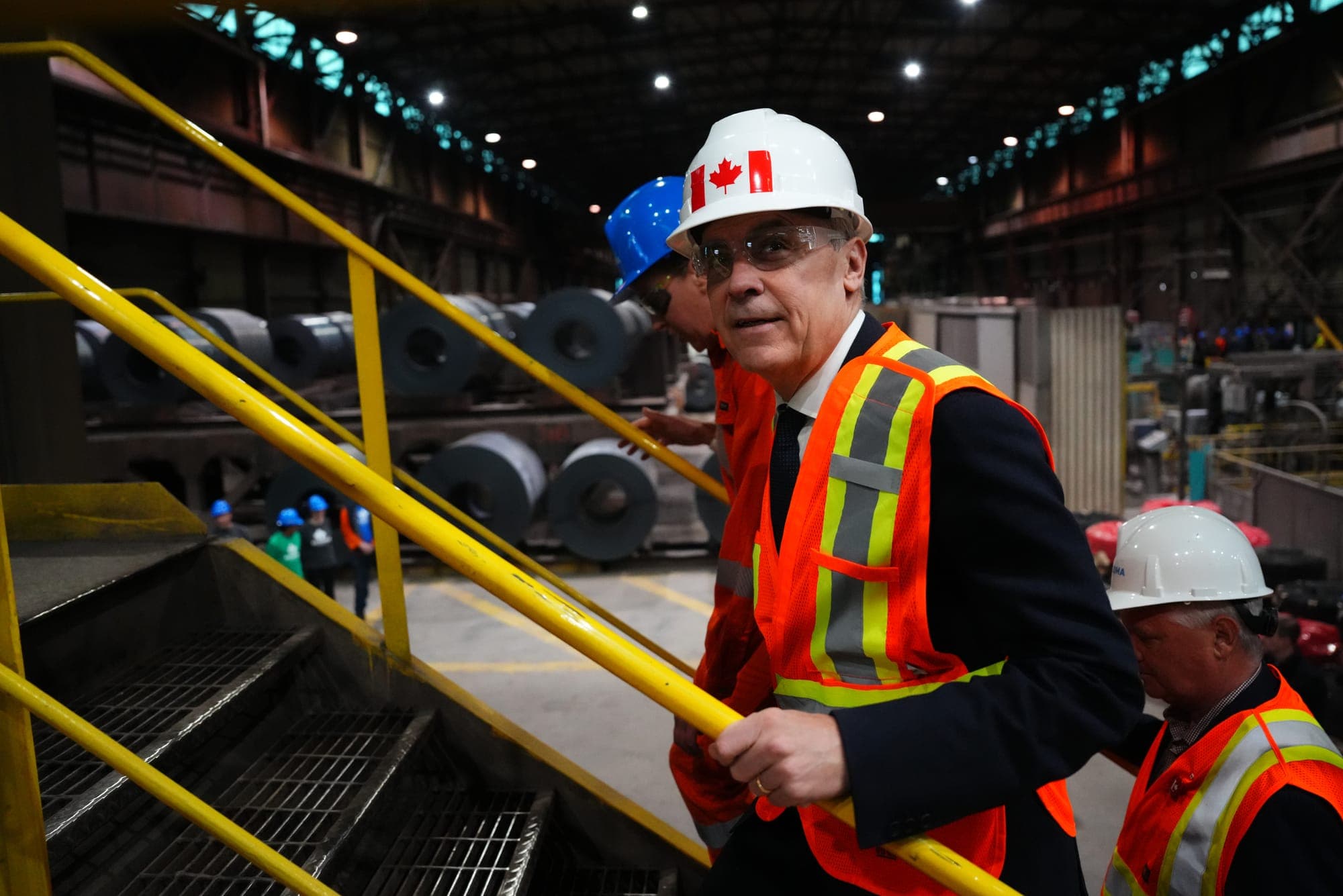 Mark Carney wears a hardhat and a high-vis orange safety vest as he climbs steps in a steel facility.