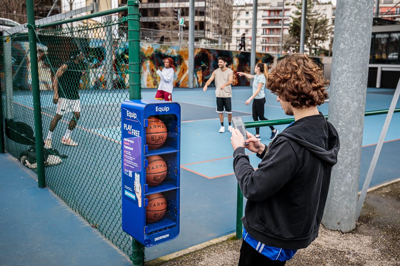 A person use an Equip Sport app on their phone to access free basketballs from a self-serve station in a public park.