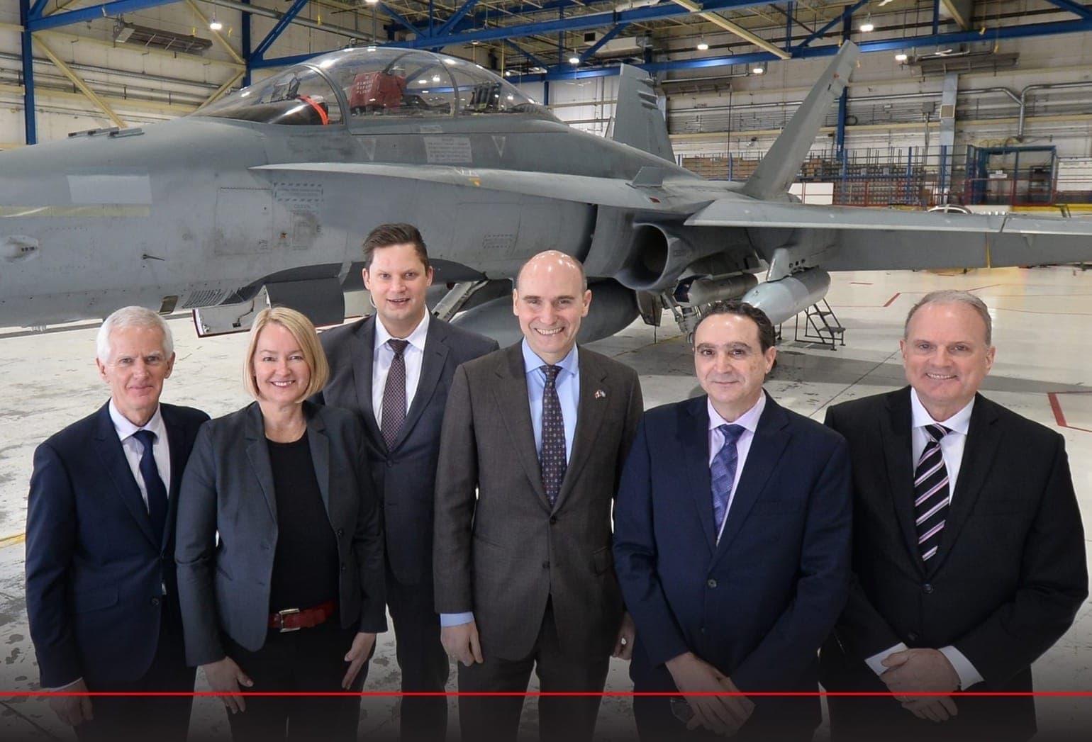 Six government officials stand in front of a fighter jet in a large warehouse at L3Harris' Mirabel, Que. facility.