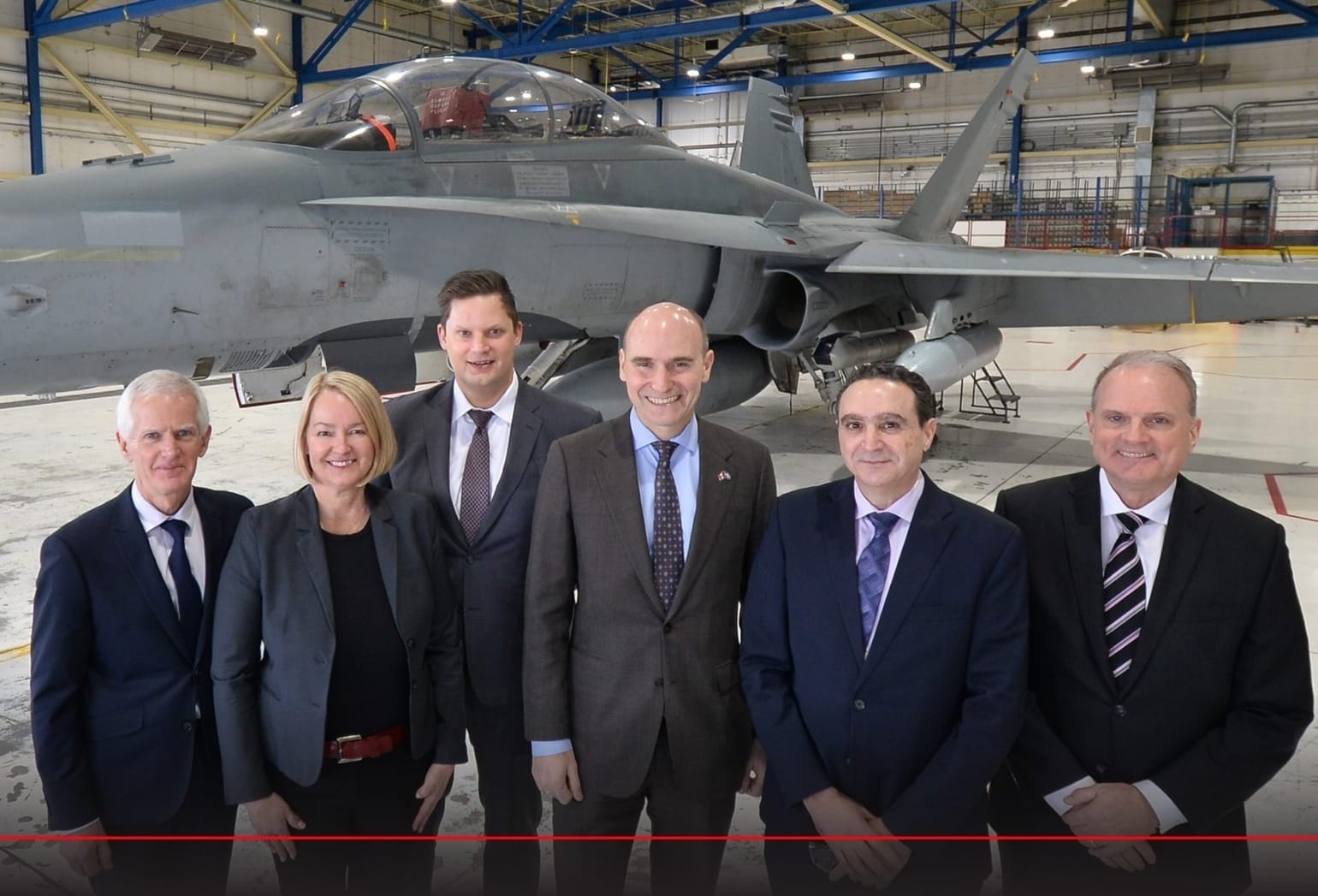 Six government officials stand in front of a fighter jet in a large warehouse at L3Harris' Mirabel, Que. facility.