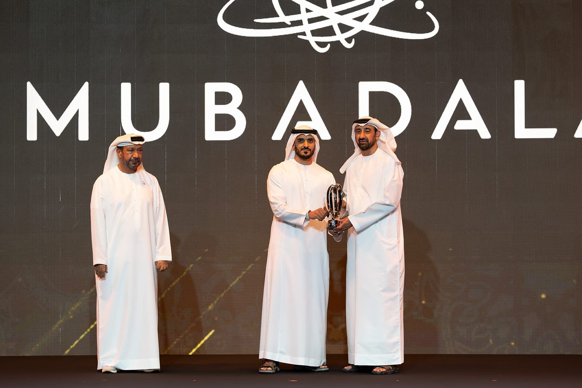 Two men in white robes shake hands as they exchange an award. They are standing in front of a backdrop that says MUBADA