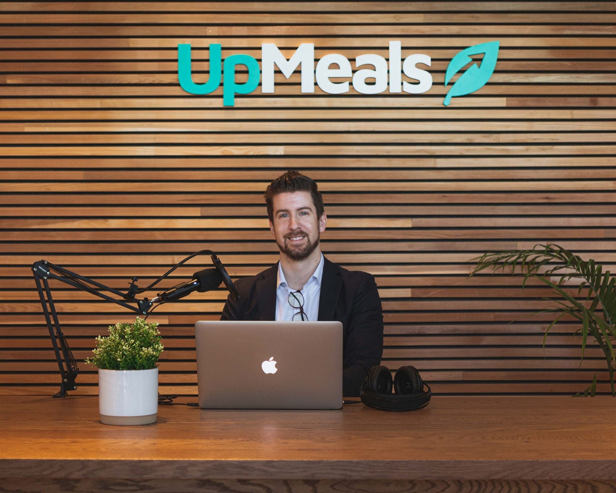 Drew Munro sits behind a microphone and a laptop in front of a wood-panelled wall reading 'UpMeals.'