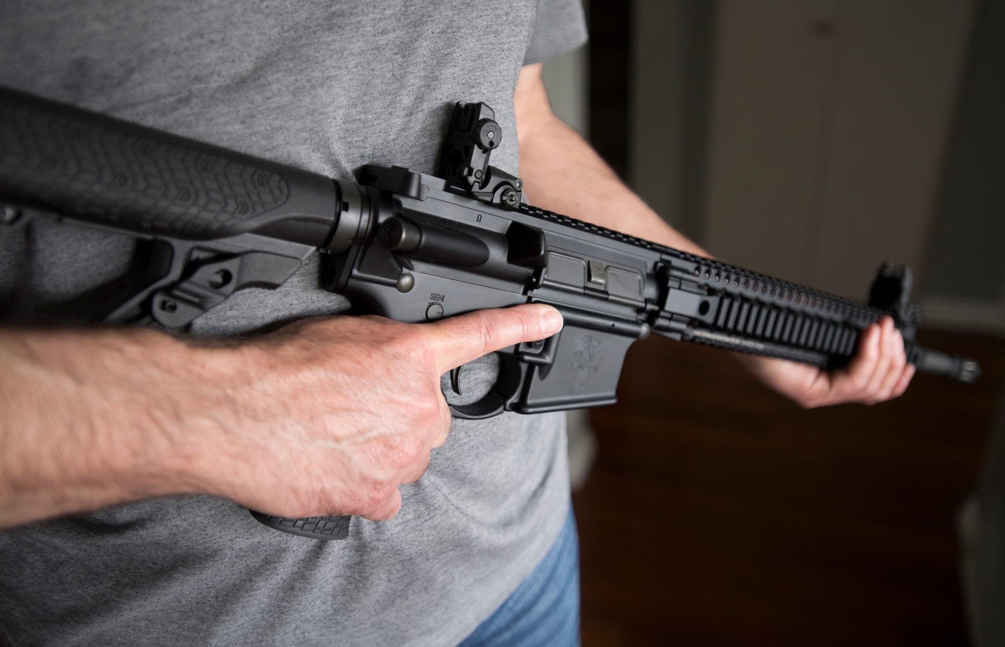 A person wearing a grey shirt and blue jeans holds a black assault rifle.