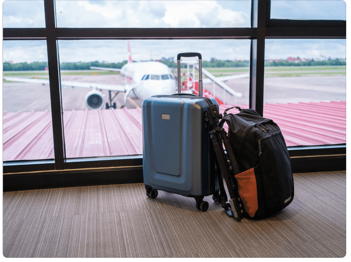 A backpack leans against a carry-on suitcase. Through a window behind the luggage, a plane can be seen parked on a tarmac.