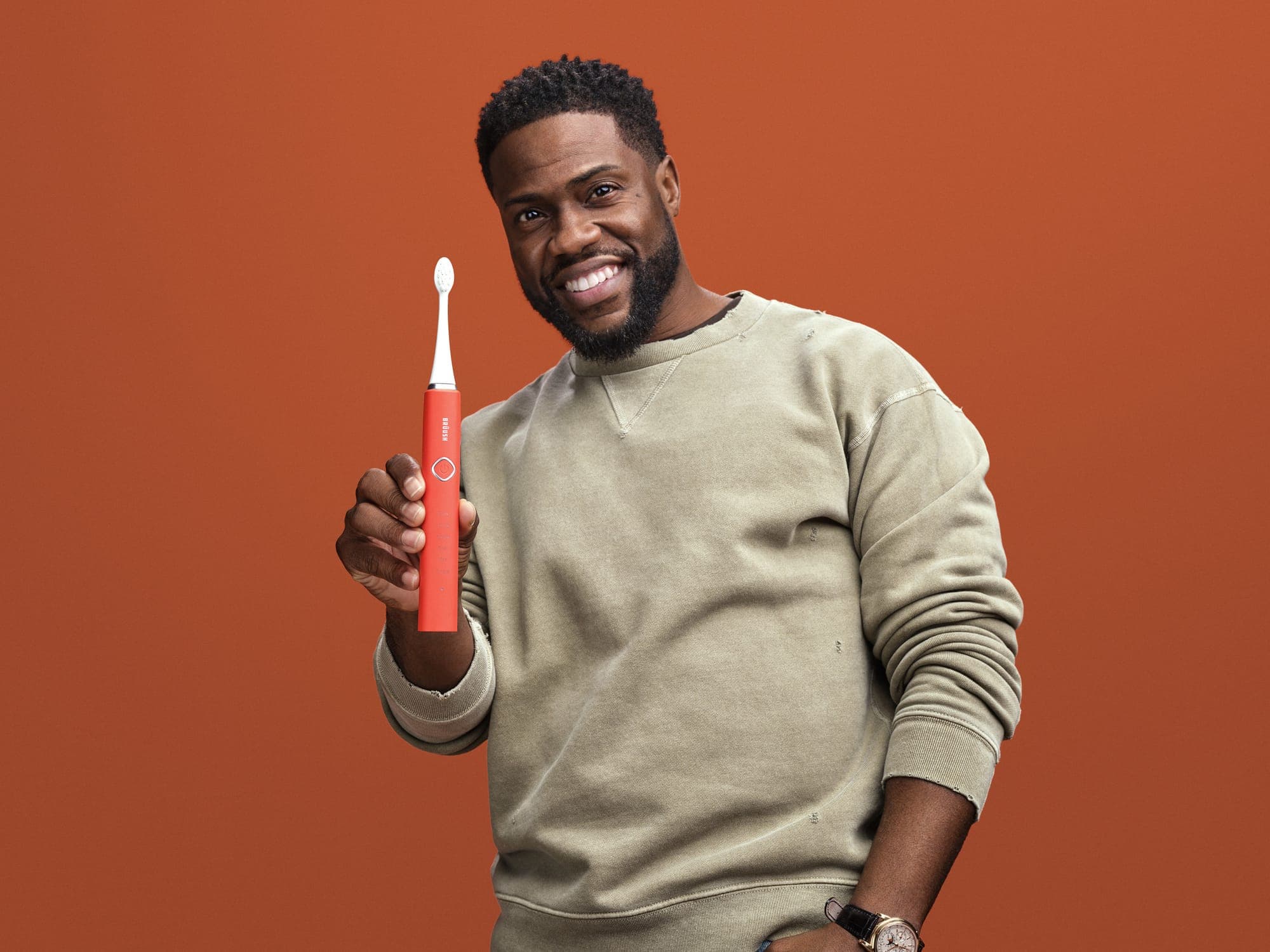 A smiling Kevin Hart is shown from the waist up, wearing a beige sweatshirt and holding an orange electric toothbrush.