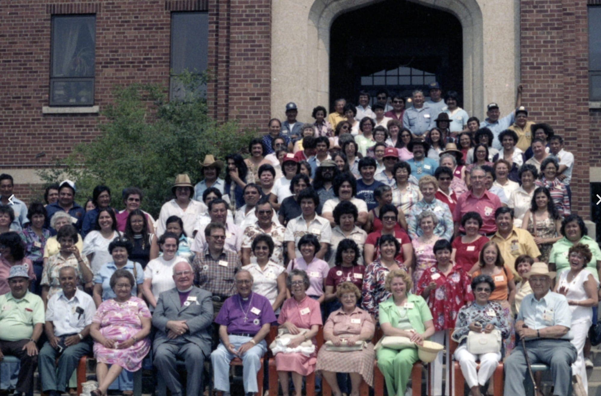 A group of survivors gather in front of what used to be a residential school building and pose in different coloured shirts
