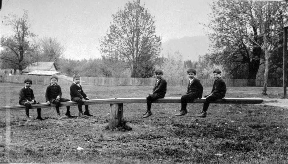 A black and white historical image shows six Indigenous boys balancing on a seesaw.