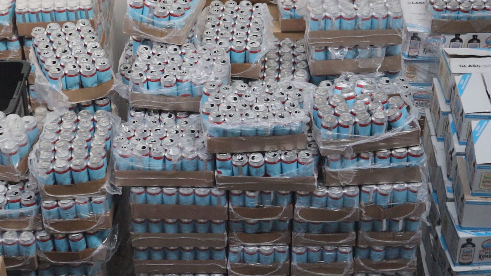 Several large pallets of beer in teal and maroon cans are pictured heaped atop each other.