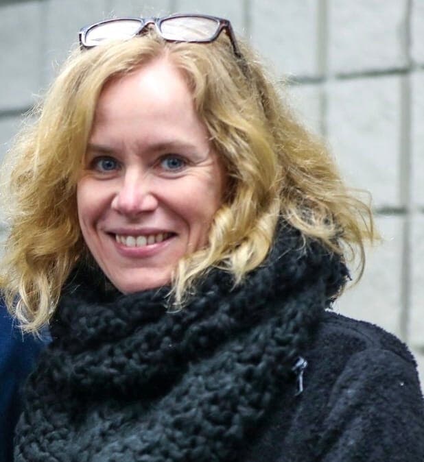 A blonde woman with reading glasses resting on top of her head, wearing a black sweater and black scarf smiles at the camera.
