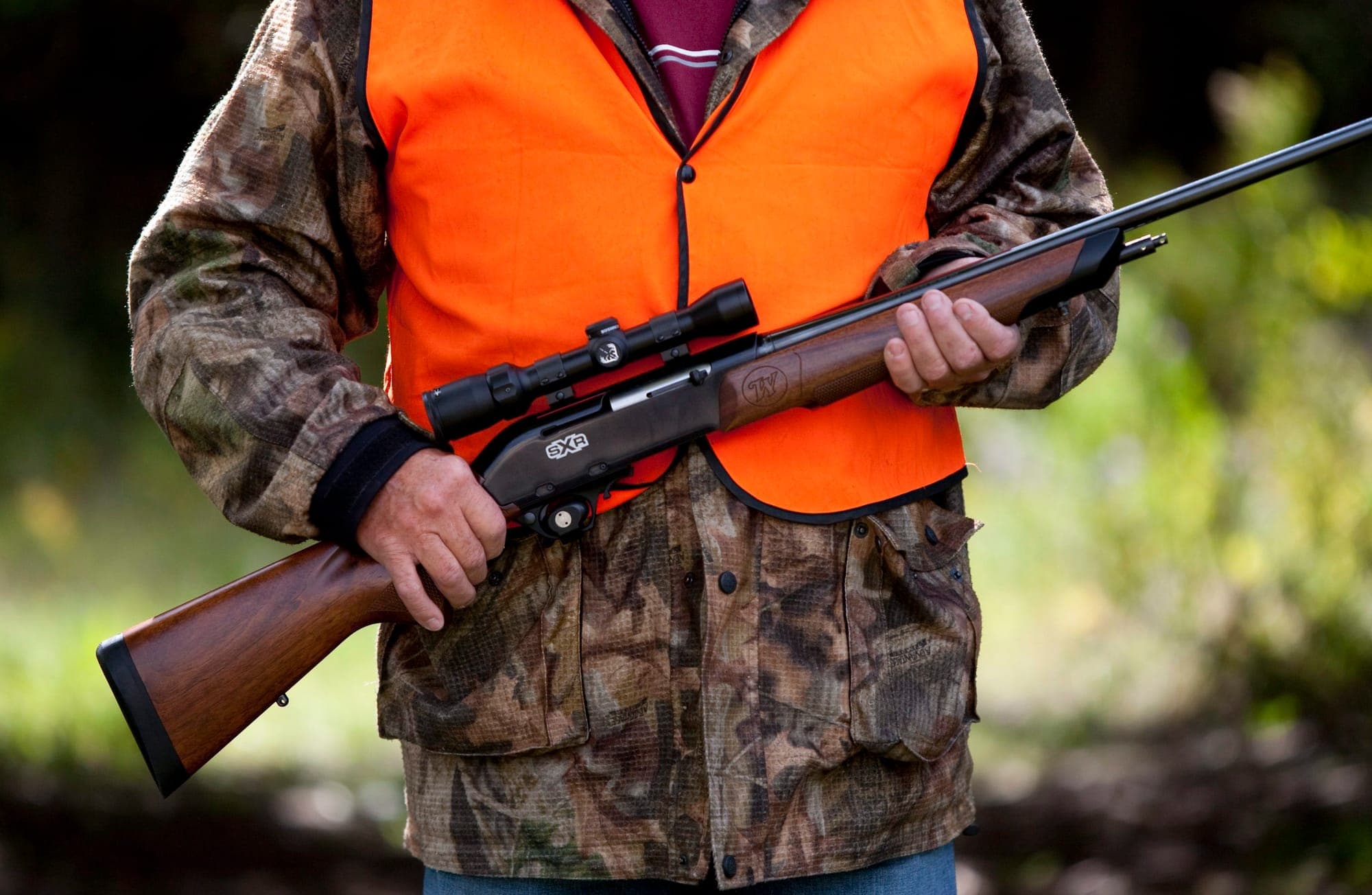 A hunter wearing a camouflage jacket beneath an orange vest holds a rifle with a scope.