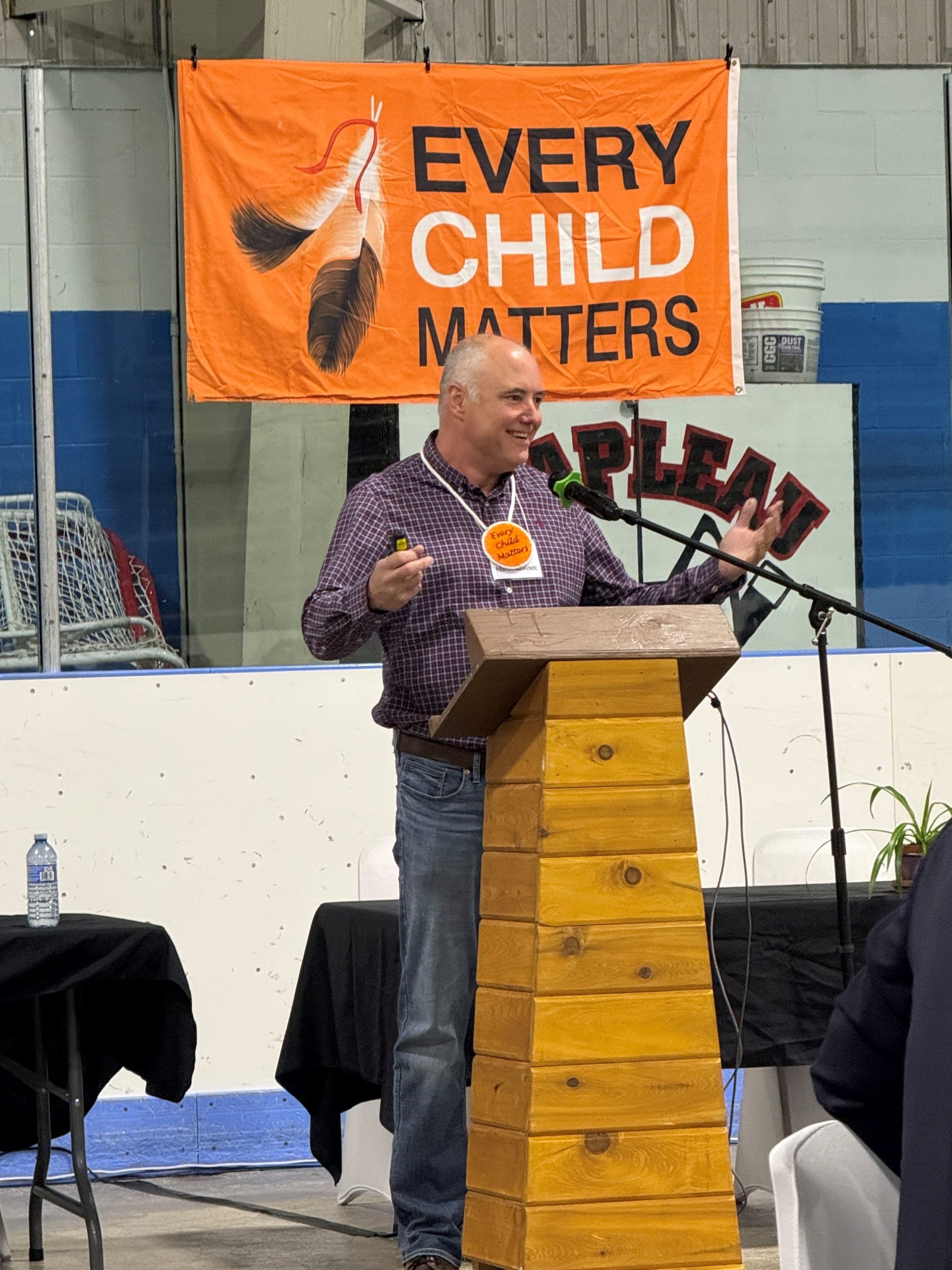 A man speaks at a podium wearing an orange beaded medallion in front of an “Every Child Matters” poster.