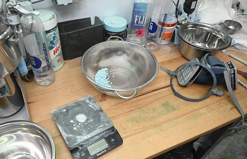 Small blue pills sit in a pile in a metal colander on top of a wooden table covered with drug production paraphrenalia.
