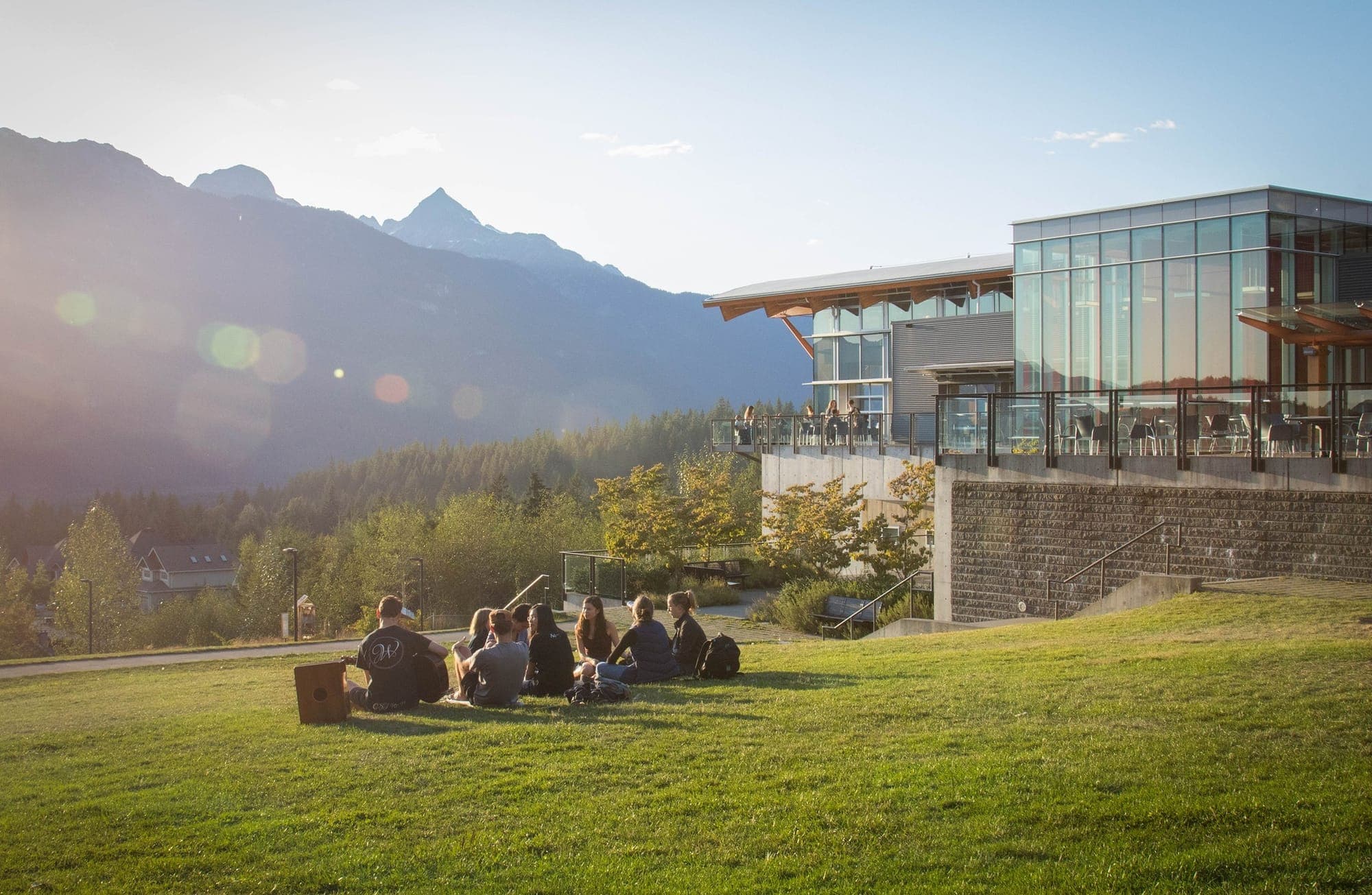 Students gather on a grassy slope below the mountains next to a glass-covered building at Quest University.