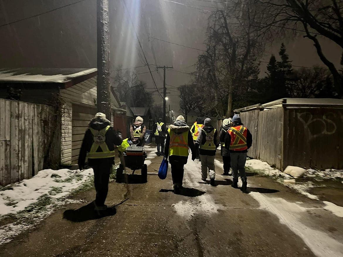 People wearing fluorescent safety vest are seen from behind as they walk through a snowy alley in the dark.