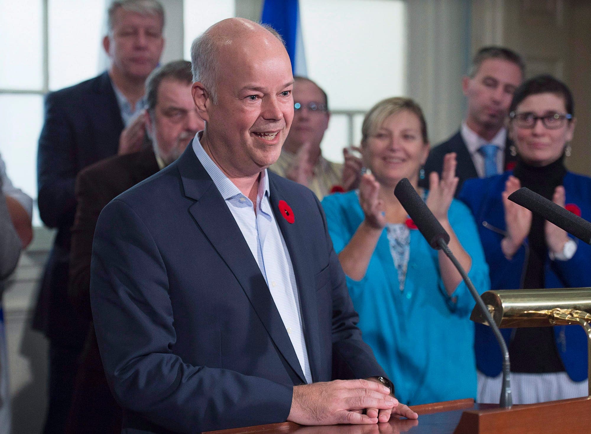 Man in blue sport jacket stands at a podium with clapping people behind him.